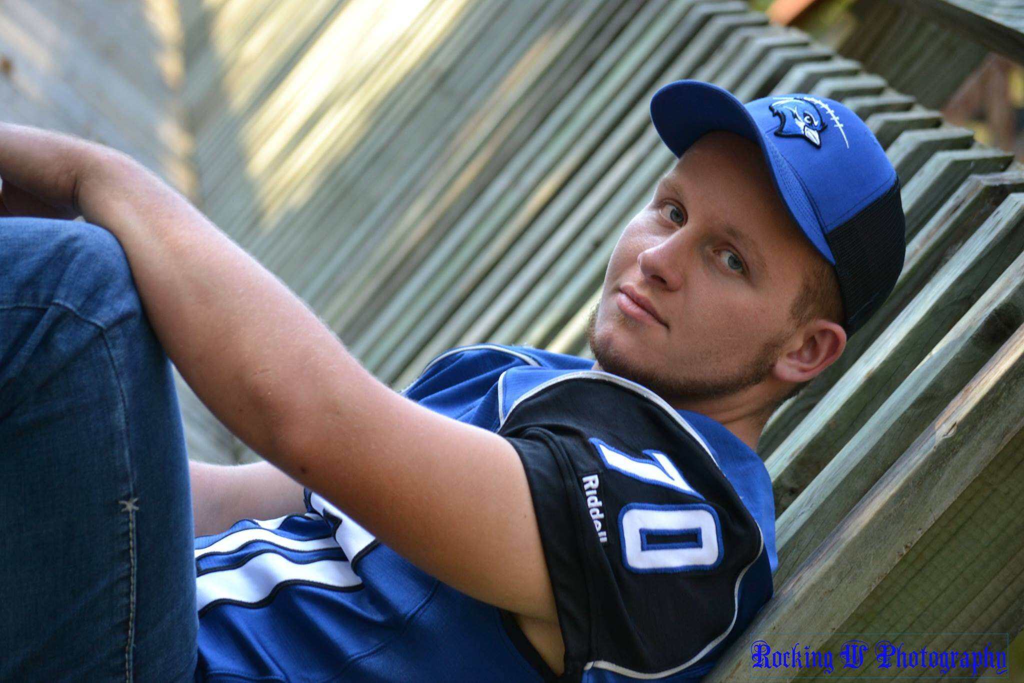 High school senior wearing a blue football jersey and cap, leaning against a wooden fence, looking confidently at the camera.